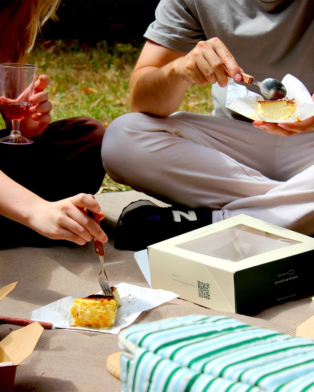Two people sharing cheesecake at a picnic, branded box with a review QR code